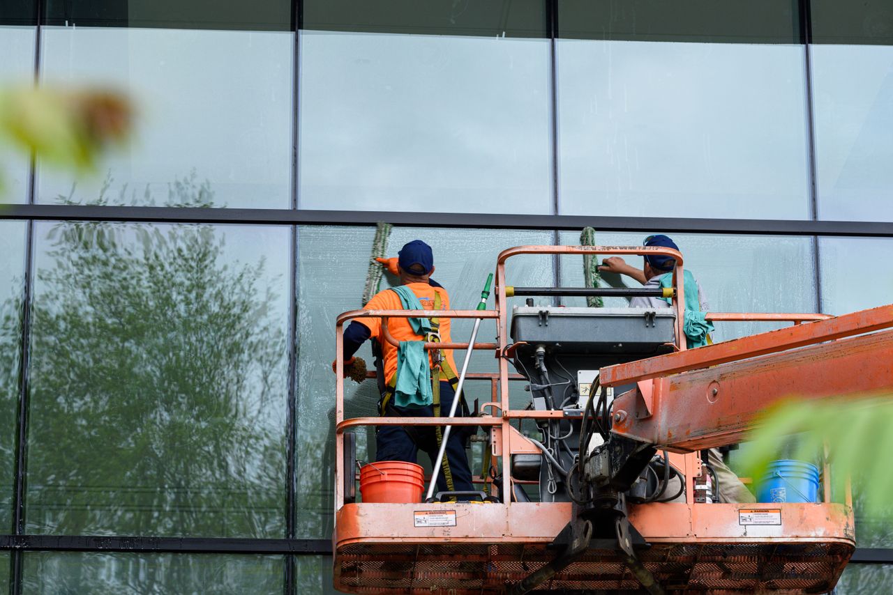 Deux travailleurs nettoient les fenêtres d’une façade de bâtiment vitré à l’aide d’une nacelle, afin d’améliorer l’apparence de l’édifice.